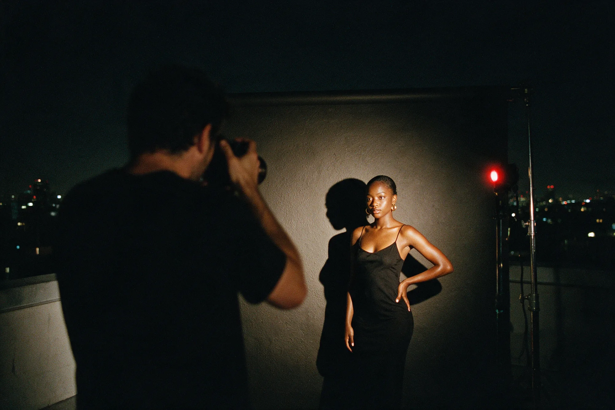 A flash firing on a night shoot: the photographer in silhouette, the creator lit hard against a rooftop wall