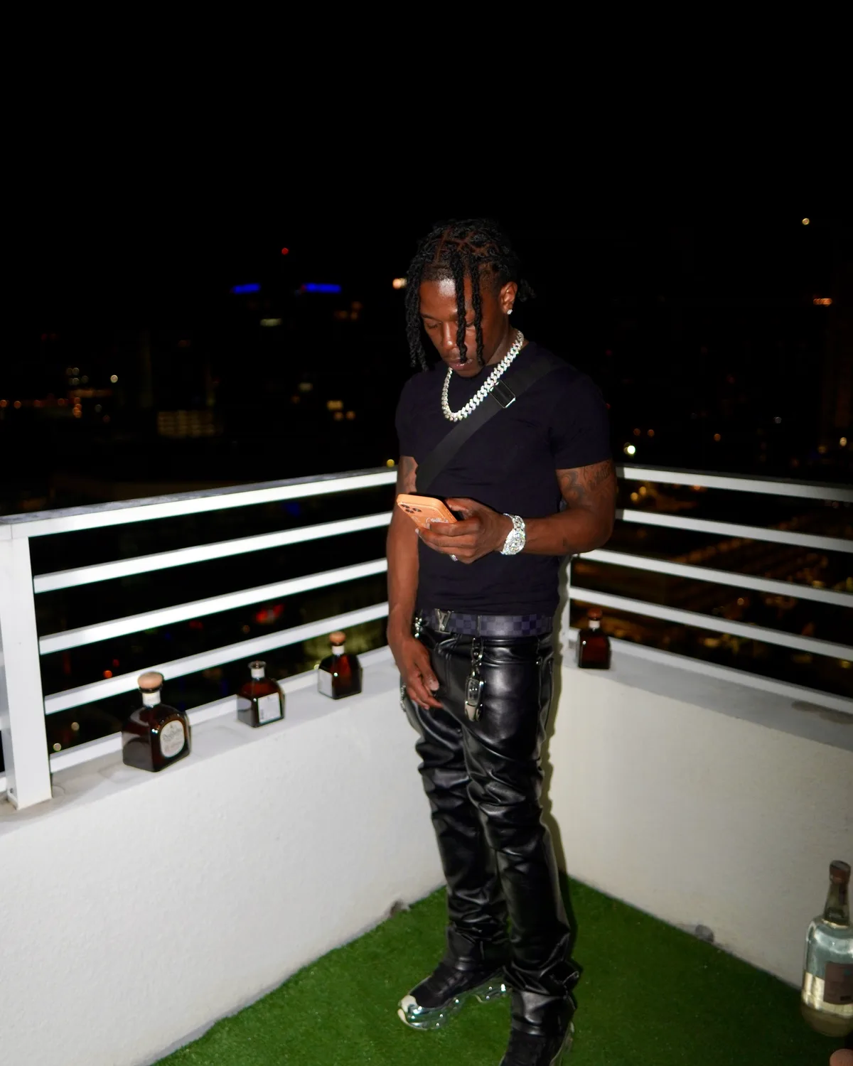 A man in a black tee and leather trousers on a rooftop at night, lit by direct flash against the city